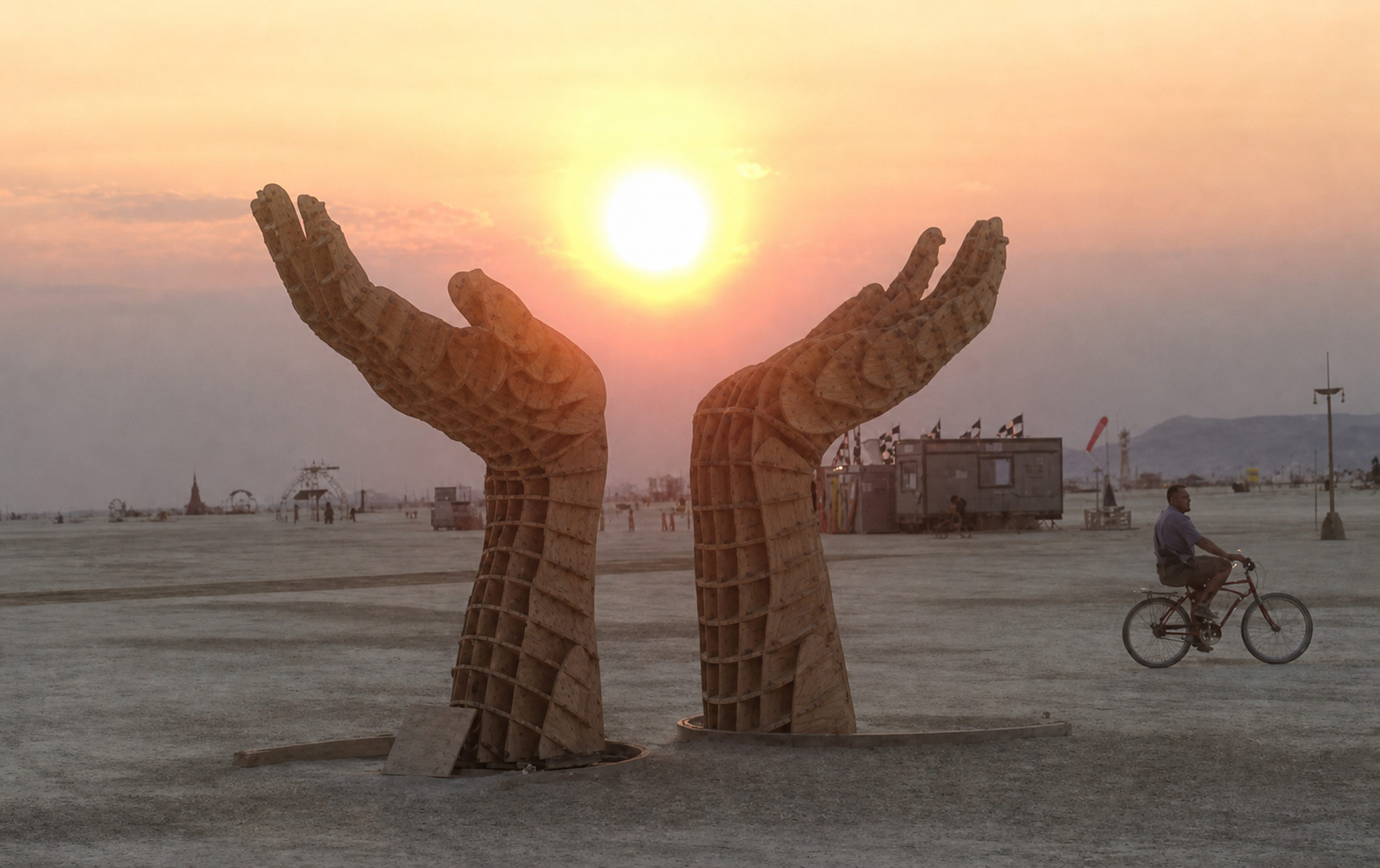 Two monumental wooden hands rising from the playa, palms open around the setting sun, a cyclist passing behind