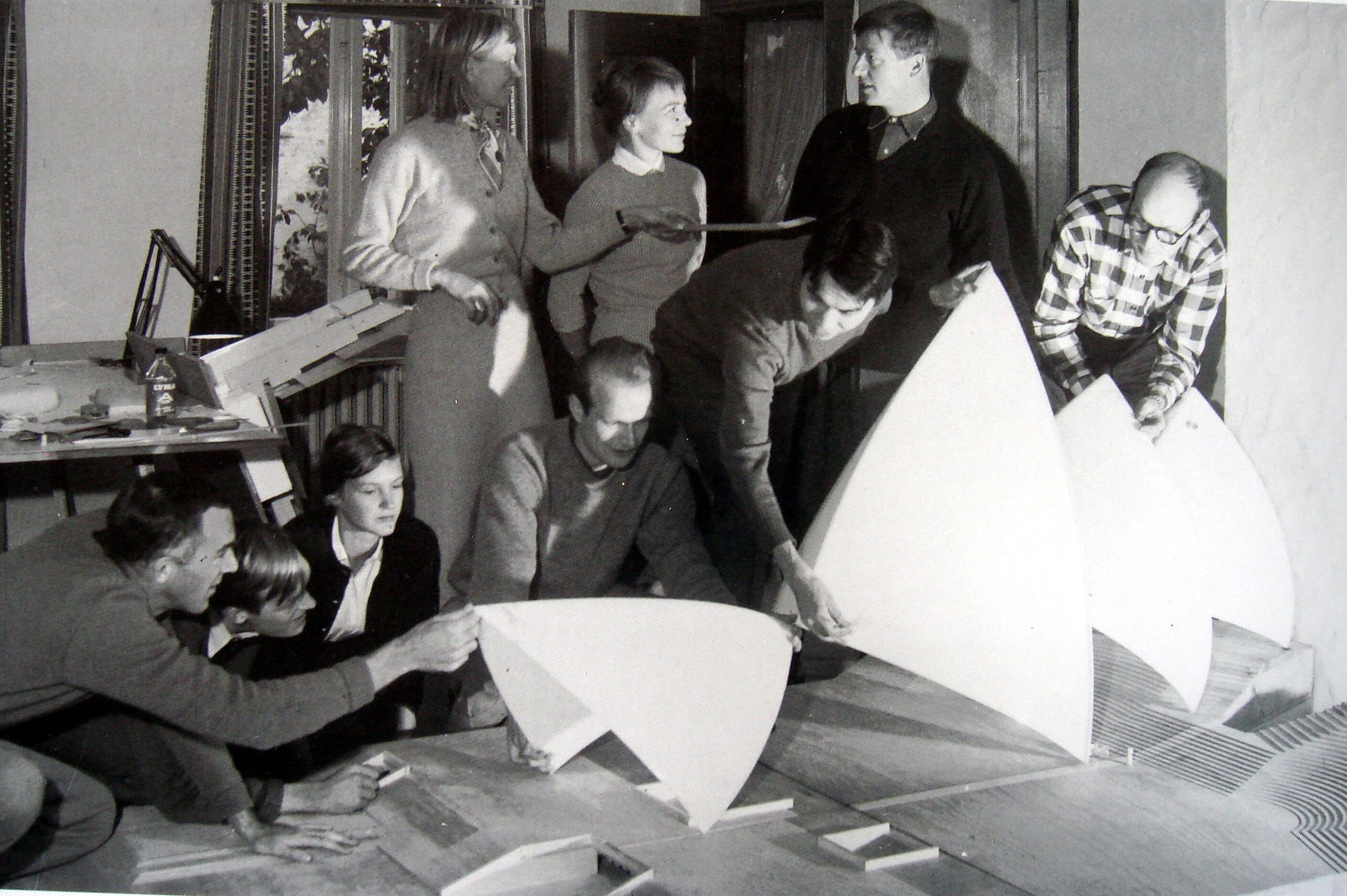 A design team gathered around large curved shell-shaped model panels on a workroom table, one member adjusting a piece while others confer